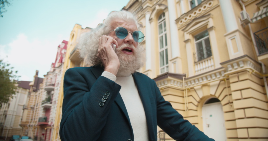 Happy old bearded man in trendy glasses talking on smartphone, smiling outdoor