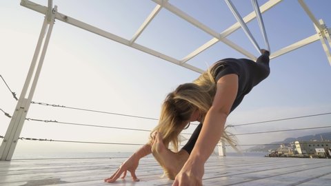 Athletic young woman in skinny tracksuit does legs split leaning on hanging sling and empty pier ground near tranquil ocean water on sunny day closeup