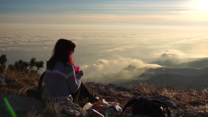 Young redhead woman in sunglases sitting on a rock high in the mountains, above the clouds and enjoying the warm sunset. Copy space. The concept of travel, rest and unity with nature | Shutterstock HD Video #1064102392