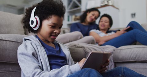 Mixed race girl using a digital tablet with headphones on. with mixed race female couple embracing on a couch. self isolation quality family time at home together during coronavirus covid 19 pandemic.
