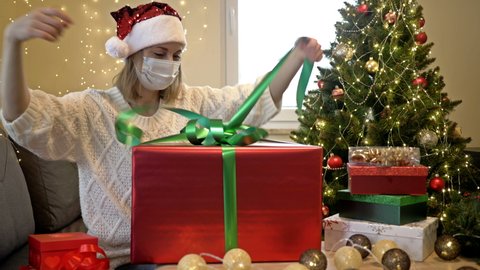 Woman in a Santa hat and a protective mask on her face is neatly unpacks his Christmas present. Against the background of the Christmas tree. Christmas despite the COVID-19 pandemic.
