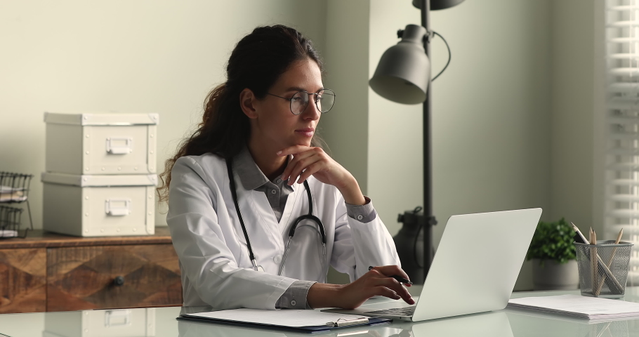 Young female doctor in white coat working on laptop in modern hospital office room, therapist typing on computer consult patient online, makes research, noting, takes useful information from internet Royalty-Free Stock Footage #1064112685