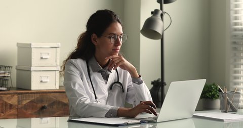 Young female doctor in white coat working on laptop in modern hospital office room, therapist typing on computer consult patient online, makes research, noting, takes useful information from internet