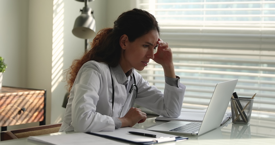 Female doctor in white uniform sit at desk read news on laptop learn information statistics of coronavirus infected patients feels stressed, problems at work, professional failure, dismissal concept | Shutterstock HD Video #1064112820