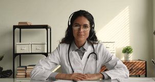 Webcam view confident female therapist in white coat sit indoor wear headset greets patient starts consultation by video call. Professional development for doctors online webinar, telemedicine concept