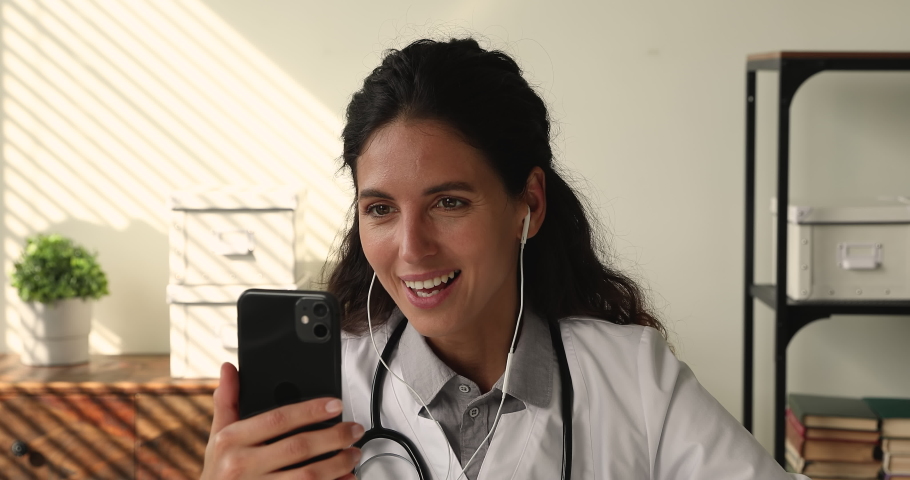 Young female medical worker gp therapist sit at workplace in clinic office wear white coat holds smartphone talk to patient using video conference app, consulting client remotely, telemedicine concept Royalty-Free Stock Footage #1064112931