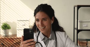Young female medical worker gp therapist sit at workplace in clinic office wear white coat holds smartphone talk to patient using video conference app, consulting client remotely, telemedicine concept