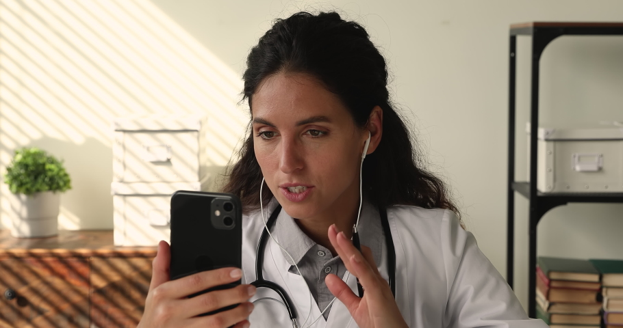Young female medical worker gp therapist sit at workplace in clinic office wear white coat holds smartphone talk to patient using video conference app, consulting client remotely, telemedicine concept | Shutterstock HD Video #1064112931