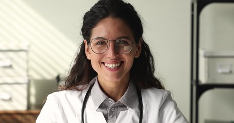 Close up view head shot beautiful 35s young physician worker wear white coat and glasses smiling looking at camera. Health professional, highly educated nurse practitioner, physician portrait concept