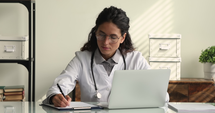 Female therapist wear white coat sit at workplace desk working on laptop holds pen makes notes texting on laptop during busy workday in clinic. Medical app usage, consulting online, research concept Royalty-Free Stock Footage #1064112973