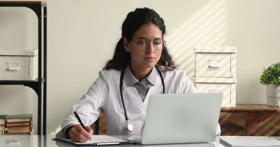 Female therapist wear white coat sit at workplace desk working on laptop holds pen makes notes texting on laptop during busy workday in clinic. Medical app usage, consulting online, research concept | Shutterstock HD Video #1064112973