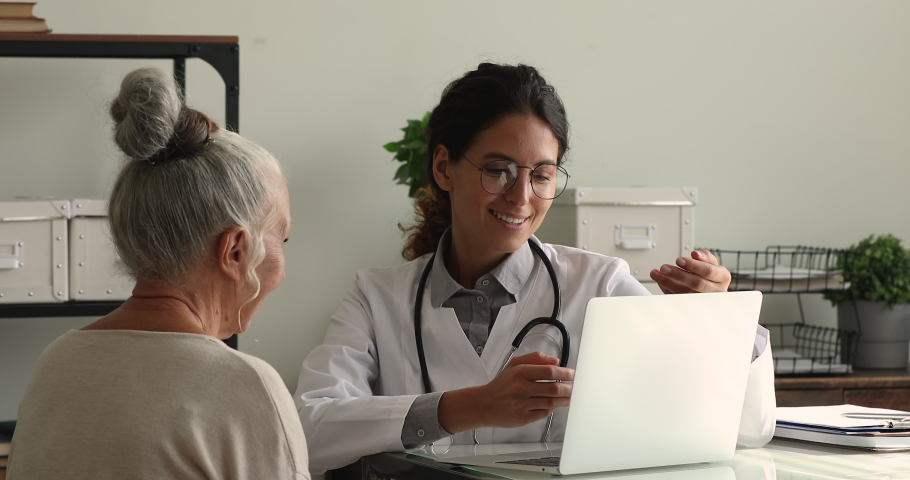 Doctor in white coat consulting to older female 65s patient seated at desk in private clinic office looking at laptop screen provide explanation about treatment disease, show medical check up results | Shutterstock HD Video #1064113012