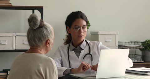 Doctor in white coat consulting to older female 65s patient seated at desk in private clinic office looking at laptop screen provide explanation about treatment disease, show medical check up results
