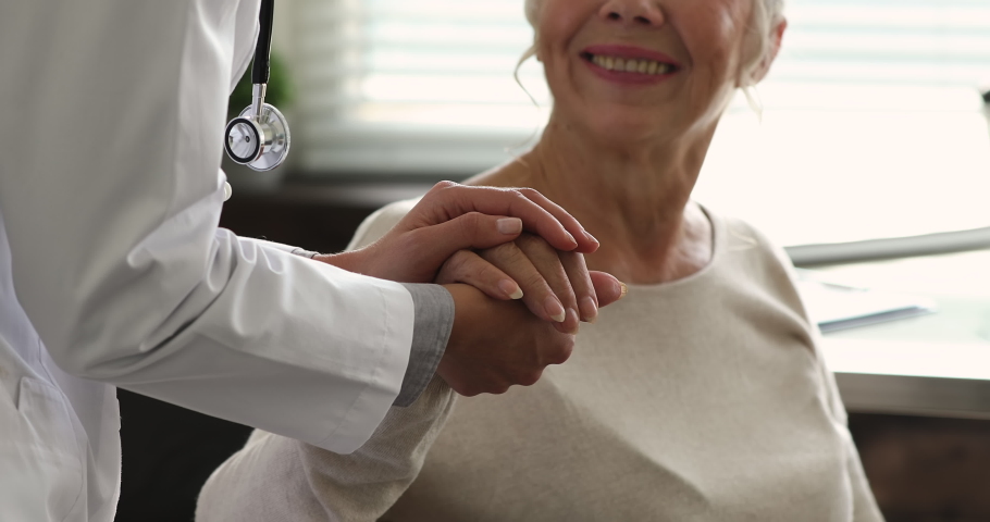 Practitioner doctor wear white coat holding hand of older patient during talk at hospital, close up. Reliable therapist provide psychological support to aged client of clinic, showing empathy concept | Shutterstock HD Video #1064113102