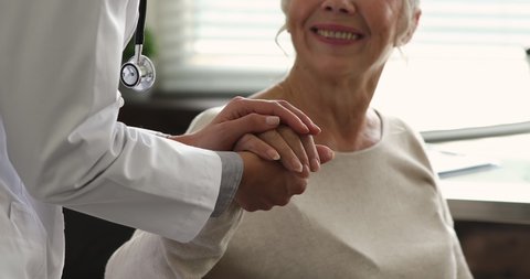 Practitioner doctor wear white coat holding hand of older patient during talk at hospital, close up. Reliable therapist provide psychological support to aged client of clinic, showing empathy concept