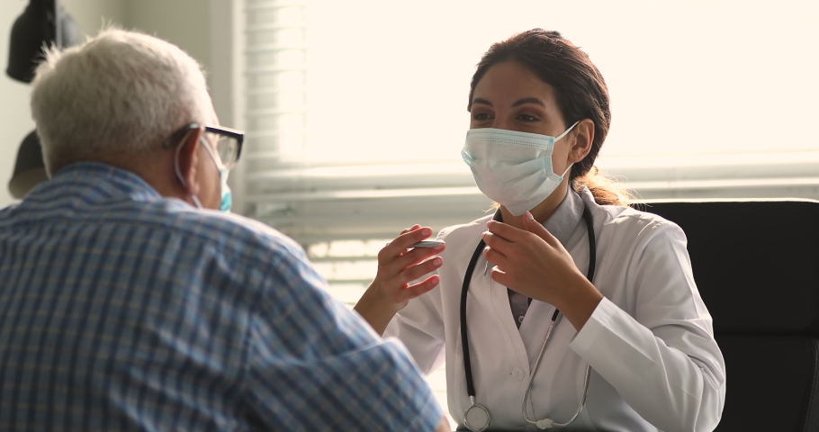 Therapist wear white coat face mask due corona virus pandemic outbreak talk to elderly patient during visit in clinic office, supporting him, telling about health check up, disease treatment concept | Shutterstock HD Video #1064113444