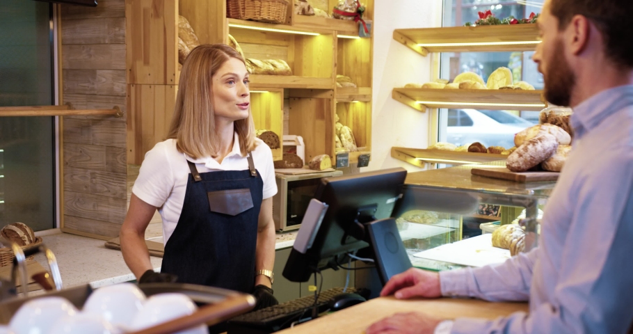 Rear of Caucasian male customer buying fresh bread in bakery shop and paying with credit card. Pretty female seller in apron selling baking in bakehouse. Small business. Shopping concept | Shutterstock HD Video #1064113603