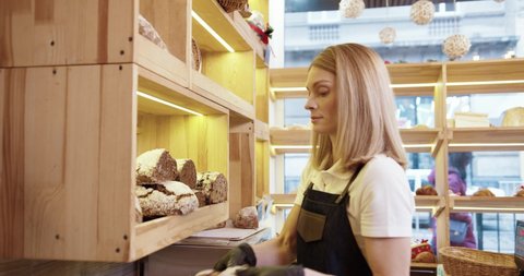 Close up of young beautiful Caucasian female seller in apron standing in bakehouse working and tputting fresh baked bread on shelves. Woman running her small own bakery shop. Food concept
