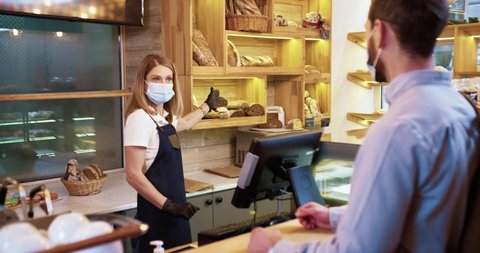 Caucasian female bakery shop worker in medical mask stands at work and speaking with male customer selling bread. Small business concept. Woman in apron at the counter in bakery shop. Food concept