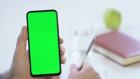 Close-up shot of green screen template smartphone in male hands at home offise, man is watcing content, makes notes in a notebook. Modent technology and information concept.
