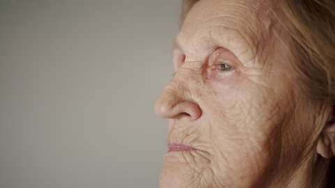 Portrait of wrinkled female face looks into distance. Serious senior woman looking out of window at home. Close up face of mature woman. Shot in 6K
