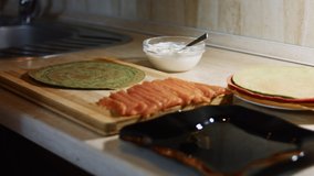 Thin tasty colorful pancakes. Close-up of woman's hand spreading Philadelphia cream cheese as filling on green pancake on a wooden board. 4K video.