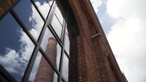 Old chimney of Moscow Arma business quarter reflected in a window. Action. Bottom view of a red brick wall of an old gas factory with a high chimney on blue cloudy sky background.