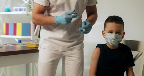 Little boy kid in protective medical mask is getting vaccinated. Unrecognizable male doctor injects vaccine into child's shoulder.
