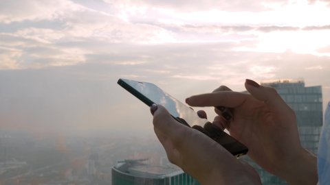 Leisure time, entertainment, sightseeing, technology concept. Close up side view - woman using smartphone device against cityscape view through window of skyscraper in office - scrolling and touching