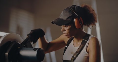 Confident black female laborer in headphones and glasses using metal cutting saw and smiling at camera folding arms in confidence