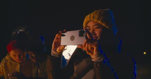 Excited Asian female in warm hat using smartphone to record glowing celebration firework standing in crowd on street