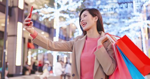 asian woman takes selfie by smart phone with some bags on her hand after shopping at night