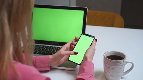 Close up woman using smartphone green screen on the table with laptop computer Chromakey. Close up shot of woman's hands holding mobile phone swiping scrolling green screen.