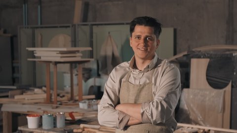 Medium close up portrait of craftsman in vintage shirt and apron standing with hands folded in cozy carpentry workshop smiling at camera