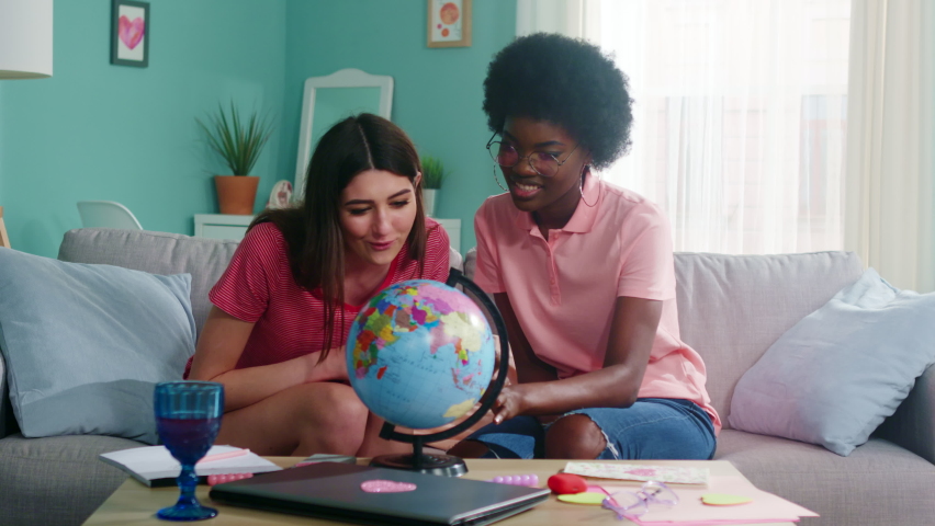 Young women of mixed races, students are sitting on sofa at home and researching the globe, working on common academic project, searching for information about countries, Foreground, Slow motion.