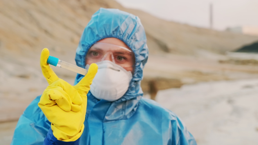 Closeup of female ecologist in protective suit, respiratory mask and yellow rubber gloves holding test tube filled with contaminated poisoned paddle water from dirty industrial zone | Shutterstock HD Video #1064139985