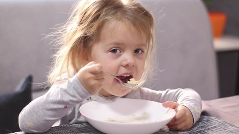 pretty little blonde girl in pajama eating milk porridge with asterisk vermicelli. breakfast, morning home. FullHD footage.
