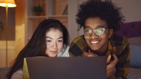 Happy diverse couple lying on bed while using laptop in evening. Smiling multiethnic man and woman relaxing in bedroom and watching movie or shopping online on computer - Powered by Shutterstock - Get 15% off with code: PIKWIZARD15
