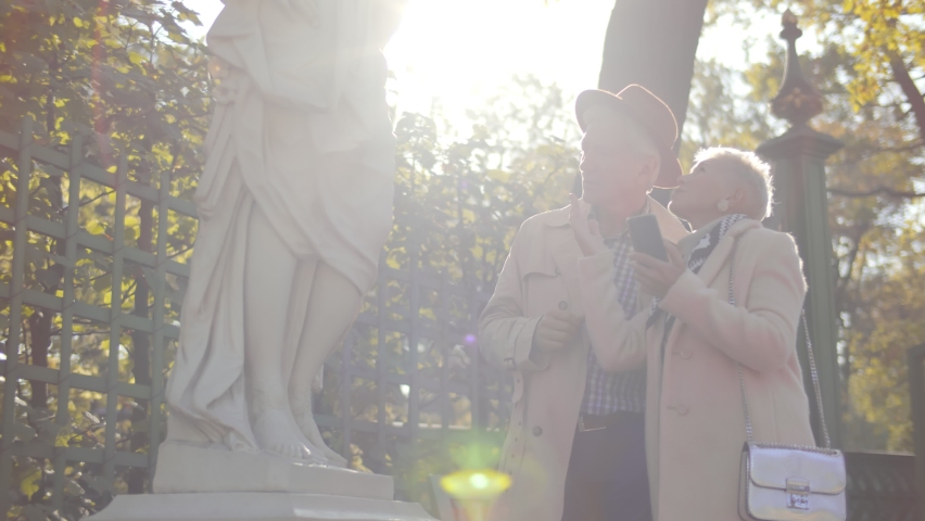 A stylish aged couple standing near a sculpture in the garden and reading information on a mobile phone. Mature man and woman tourists walking in city park. Autumn vibes.
