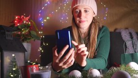 Young woman in hat holds smart phone make video call on Christmas Eve. Congratulates friends relatives and show present for them