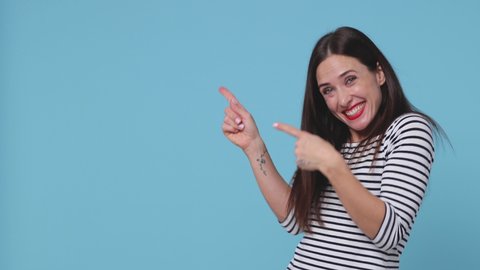 Cheerful excited brunette young woman 20s wearing striped shirt posing isolated on blue background studio. People lifestyle concept. Pointing index finger aside camera showing thumbs up winner gesture