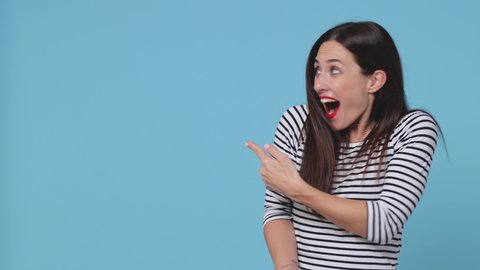 Amazed surprised brunette young woman 20s wearing basic striped shirt posing isolated on blue color background studio. People lifestyle concept. Pointing index finger aside showing thumbs up say wow
