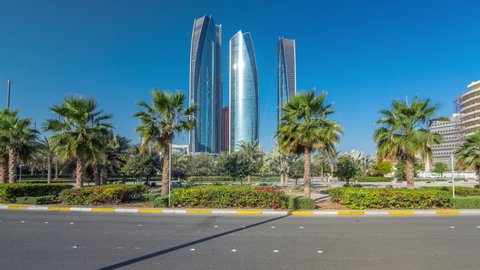 Skyscrapers of Abu Dhabi at morning with towers buildings timelapse hyperlapse. Park with palms and traffic on the road. Sun reflected from glass surface. United Arab Emirates