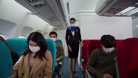 Beautiful Asian female flight attendant walking along aisle checking the multiracial passengers on safety standards before take off. Everyone on the plane wearing a face mask for protective Covid-19. 