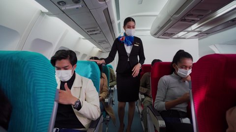Beautiful Asian female flight attendant walking along aisle checking the multiracial passengers on safety standards before take off. Everyone on the plane wearing a face mask for protective Covid-19. 
