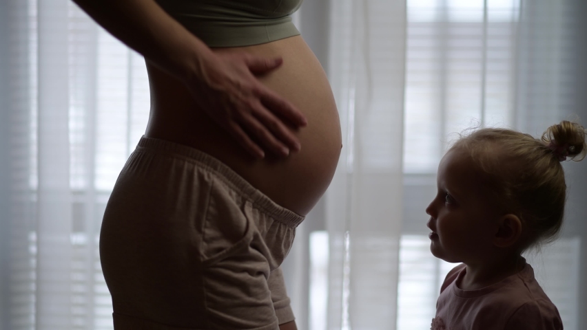 Little girl kissing her young mom's pregnant belly on a background of a window. Image of pregnancy and expectation of a child Royalty-Free Stock Footage #1064167327