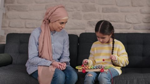 Little girl sitting on sofa at home, having fun with her mother in hijab by playing xylophone.