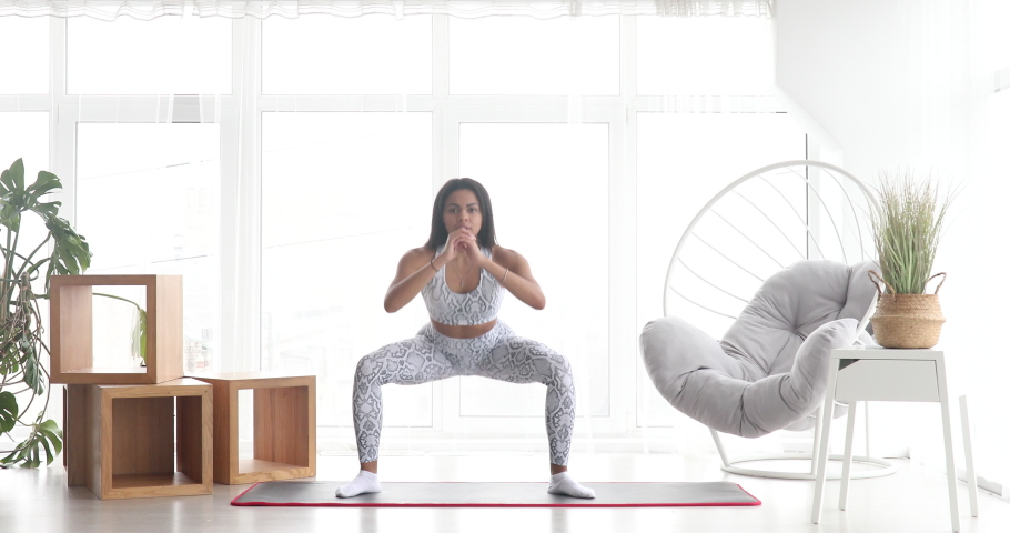 Sporty young black woman making sumo squats training at home. Athletic fit girl doing fitness aerobic exercises for booty in living room. Home fitness and wellness concept. | Shutterstock HD Video #1064169544