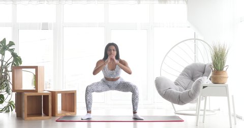 Sporty young black woman making sumo squats training at home. Athletic fit girl doing fitness aerobic exercises for booty in living room. Home fitness and wellness concept.