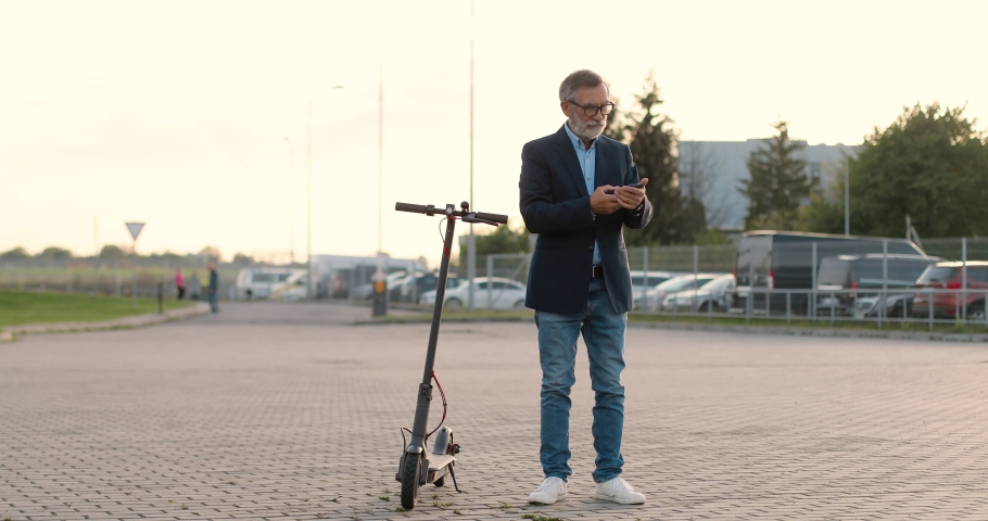 Senior gray-haired man in glasses standing at electric scooter in town and talking on mobile phone. Old grandfather speaking on cellphone outdoor. Telephone call at street. Conversation.